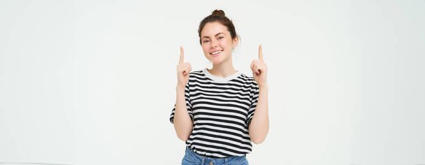 Életmód és reklám koncepció. Fiatal mosolygós nő, modern lány, ujjal mutogatva promo, mutatja banner, hely szöveges, elszigetelt fehér háttér. - Fotó, kép
