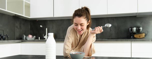 Πορτρέτο της νεαρής όμορφης γυναίκας με μπουρνούζι, να τρώει δημητριακά για πρωινό, γέρνει στον πάγκο της κουζίνας, κοιτάζοντας το πρωινό της γεύμα. - Φωτογραφία, εικόνα