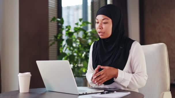 Líder de equipe afro-americano confiante sentado na frente de um computador portátil em um escritório acolhedor e falando online. Recrutadora de meia-idade em hijab preto conversando com buscador durante entrevista em vídeo. - Filmagem, Vídeo