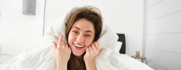 Portrait of beautiful brunette woman lying in bed, resting in her hotel room, covered in white blankets, laughing and smiling at camera. - Photo, Image