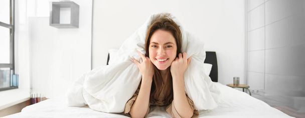 Słodka dziewczyna z brudne włosy, leżące w łóżku pokryte białymi prześcieradłami kołdrę, uśmiechając się i śmiejąc coquettish, spędzając czas w jej sypialni. - Zdjęcie, obraz