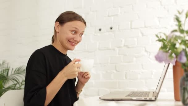Усміхнена молода жінка фрілансер розмовляє і махає рукою привіт під час онлайн відеодзвінків через ноутбук, сидячи в кав'ярні з чашкою кави. Бізнес-леді проводить конференцію в Інтернеті через ПК - Кадри, відео