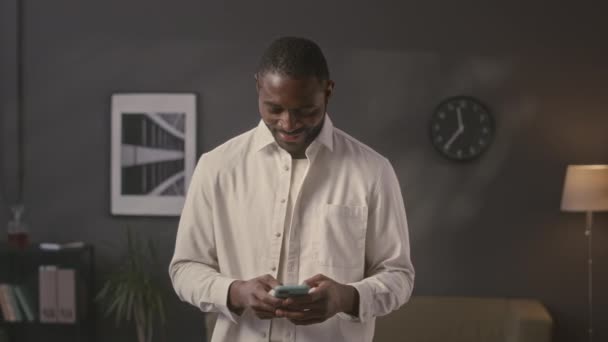 スマートフォンでブラウジングし,ミニマリストのオフィスに立っているカメラのためにポーズする若いアフリカ系アメリカ人のビジネスマンの中型肖像画 - 映像、動画