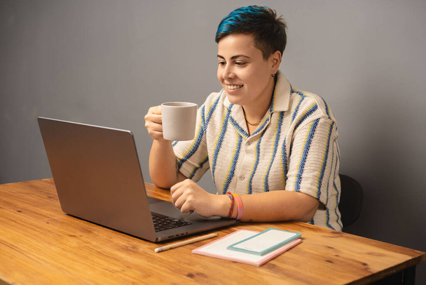 Nowoczesna i stylowa kobieta o niebieskich włosach siedzi w swoim biurze, sącząc kawę i pracując nad laptopem, prezentując współczesną oprawę pracy z domu. - Zdjęcie, obraz
