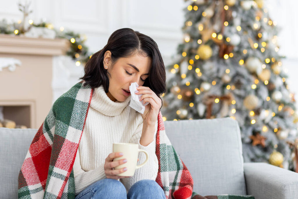Una giovane donna si ammalò durante le vacanze di Natale e Capodanno. Si siede a casa sul divano coperto da una coperta, si asciuga il naso da un naso che cola con un tovagliolo, tiene una tazza di bevande, medicine. - Foto, immagini