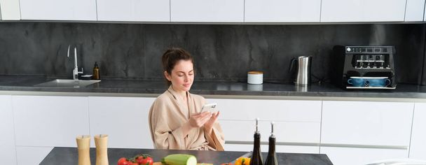 Portret van een vrouw in badjas zittend in de keuken met smartphone, koken diner, kijken naar recept op sociale media, video tutorial hoe om te koken. - Foto, afbeelding