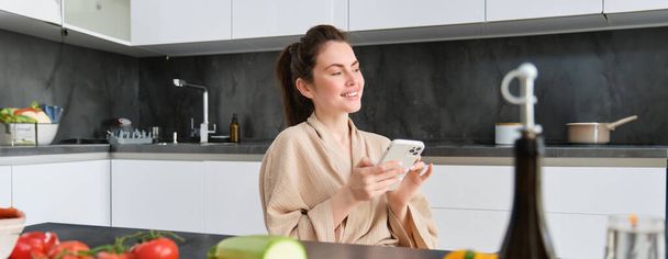 Ritratto di bella giovane donna con smartphone, seduta in cucina, cena di cucina, verdure sul tagliere. Ragazza guardando ricetta di cucina sul telefono cellulare. - Foto, immagini