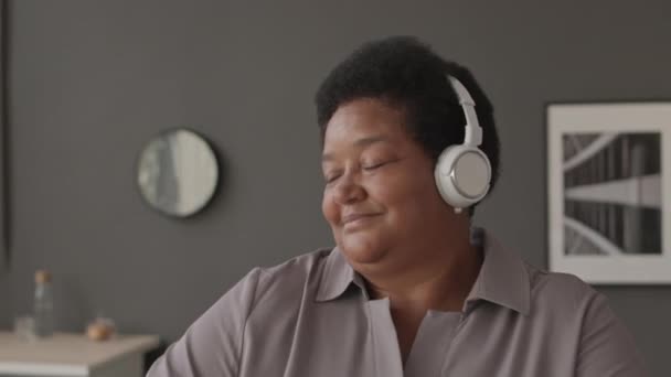 Brust hoch für eine fröhliche Afroamerikanerin mittleren Alters, die in drahtlosen Kopfhörern energiegeladene Musik hört und während der Arbeitspause am Arbeitsplatz tanzt - Filmmaterial, Video