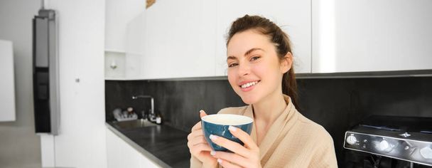 Portrait de femme moderne heureuse et souriante, commence sa journée avec une tasse de thé du matin. Fille boire du café dans la cuisine, debout avec une tasse et l'air détendu. - Photo, image