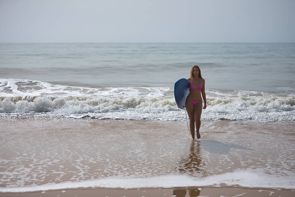 Genç, güzel, sarışın bir kadın, pembe bir bikini giyiyor, kolunun altında mavi bir sörf tahtası var, dalgalarda sörf yaptıktan sonra sudan çıkıyor. Kumsal, bikini, sörf, dalgalar, spor. - Fotoğraf, Görsel