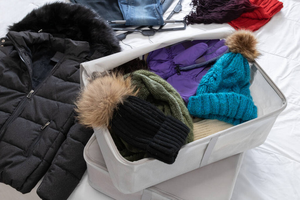 Горизонтальное фото, зимняя одежда на кровати, шерстяные шляпы и шарф, заправленные в суконные коробки, которые должны быть собраны к началу весны. Изменение шкафа. - Фото, изображение