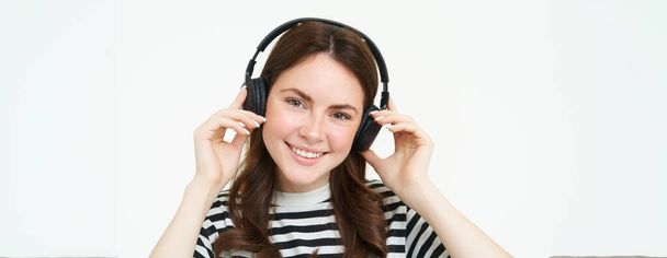 Close up retrato de linda jovem, sorrindo, colocando fones de ouvido, ouvir música em fones de ouvido, experimentando novos fones de ouvido, fundo branco. - Foto, Imagem