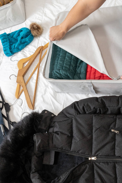 Foto verticale, mano della donna che chiude la cerniera di una scatola di stoffa, con cappotti all'interno, su un letto, raccogliendo vestiti invernali al cambio di stagione. - Foto, immagini