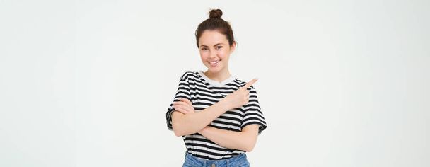 Portrait of smiling, beautiful young brunette woman, pointing finger left, showing advertisement, white studio background. - Photo, Image