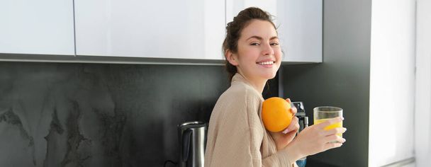 Beautiful young woman in bathrobe, drinking homemade orange juice, smiling and laughing, standing near worktop in kitchen. - Photo, Image