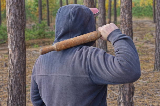 Ένας επιθετικός άντρας με μπλε ρούχα και κουκούλα κουνάει ένα καφέ ραβδί με ένα ρόπαλο πάνω από το κεφάλι του στο δρόμο ανάμεσα στα δέντρα στο δάσος. - Φωτογραφία, εικόνα