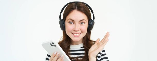 Retrato de bela jovem, usando fones de ouvido, segurando smartphone, sorrindo e parecendo feliz, isolado no fundo branco. - Foto, Imagem