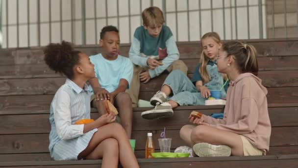 Grupo multiétnico de alunos alegres sentados em escadas ao ar livre almoçando e conversando - Filmagem, Vídeo