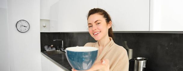 Egy csinos fiatal nő portréja ad egy csésze kávét, bögrével kínálja, a konyhában áll, fürdőköpenyben.. - Fotó, kép