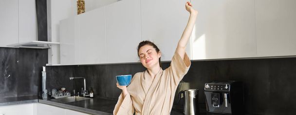 Portret beztroskiej brunetki, taniec i wyciąganie rąk w kuchni, picie kawy, uśmiech zadowolony rano. - Zdjęcie, obraz