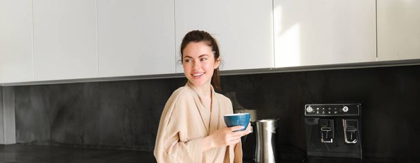 Portret pięknej gospodyni domowej, młoda kobieta w szlafroku, trzymanie filiżanki kawy, picie herbaty w kuchni, delektowanie się rankiem. - Zdjęcie, obraz