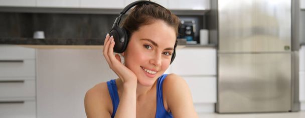 Κοντινό πλάνο πορτρέτο της όμορφης μελαχρινής κοπέλας στα αθλητικά, κάθεται σε ασύρματα ακουστικά και ακούει μουσική, ενώ κάνει προπόνηση στο σπίτι. - Φωτογραφία, εικόνα