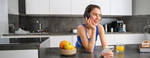 Imagen de la joven mujer deportiva feliz y elegante, parada en la cocina y bebiendo jugo de naranja, escuchando música en los auriculares, utilizando la aplicación para teléfonos inteligentes. - Foto, imagen