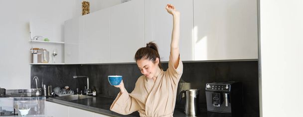 Portrait de fille heureuse dansant avec du café dans la cuisine, portant un peignoir, appréciant sa routine matinale. - Photo, image