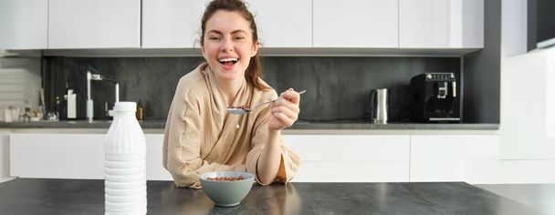 Bornozlu, güzel ve sağlıklı bir kadının portresi kahvaltısını mutfakta yiyor, mısır gevreğini sütle yiyor ve gülümsüyor.. - Fotoğraf, Görsel