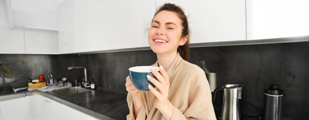 Bella giovane donna gode la sua tazza di caffè del mattino, bevendo tè dalla tazza in cucina, indossando accappatoio e sorridendo alla macchina fotografica. - Foto, immagini