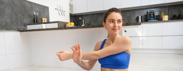 Schöne und fitte Frau trainiert zu Hause, streckt die Arme, macht Aufwärmübungen vor dem Training, trägt SportbH, sitzt auf Gummi-Yogamatte. - Foto, Bild