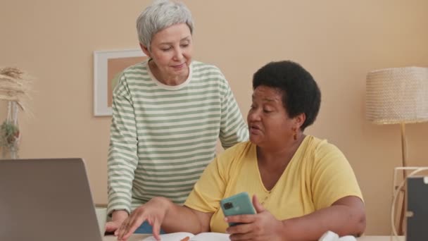 Plan moyen de deux amies âgées multiethniques utilisant un ordinateur portable et bavardant dans un appartement lumineux - Séquence, vidéo