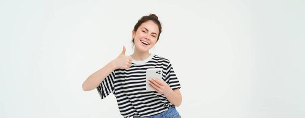 Menina entusiasmada com smartphone mostra polegares para cima, isolado sobre fundo branco. - Foto, Imagem