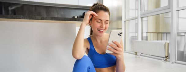 Wizerunek młodej, sprawnej kobiety noszącej sportowy stanik i niebieskie legginsy, korzystającej z aplikacji na smartfony, ćwiczącej w domu, siedzącej na podłodze i śledzącej wideo treningowe online. - Zdjęcie, obraz