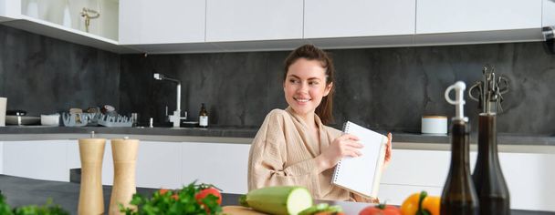 Muotokuva nainen kirjoittaa luettelon ostoksia, tehdä muistiinpanoja resepti, istuu keittiössä lähellä vihanneksia, valmistelee illallinen menu. - Valokuva, kuva