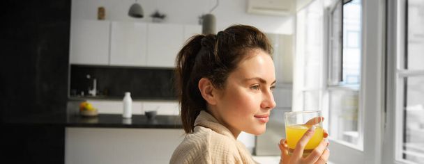 Retrato de hermosa, mujer joven moderna se despierta por la mañana, tiene su vaso fresco de jugo de naranja, mira fuera de la ventana, relajarse en el albornoz en casa. - Foto, imagen