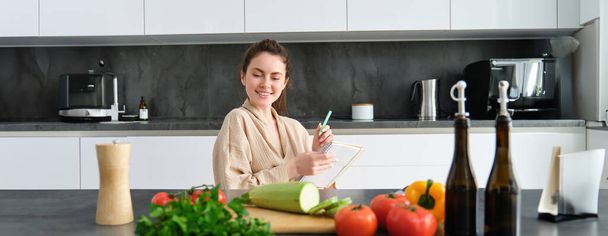 Retrato de mulher sentada com mantimentos, fazendo lista para fazer compras, posando perto de legumes, preparando comida, cozinhando na cozinha. - Foto, Imagem
