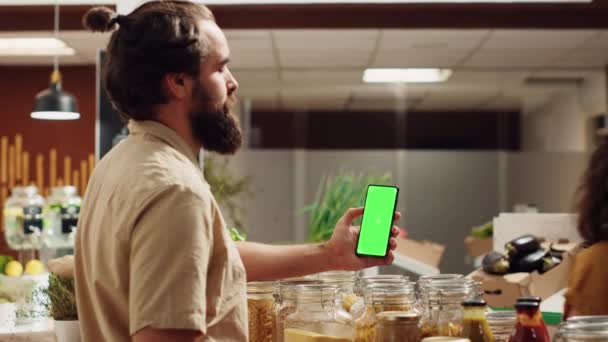 Hombre usando el teléfono inteligente de pantalla verde en el supermercado de cero residuos para comprobar los ingredientes para una receta saludable. Cliente en la tienda de comestibles local utiliza el teléfono clave de croma mientras que las compras de verduras orgánicas - Metraje, vídeo