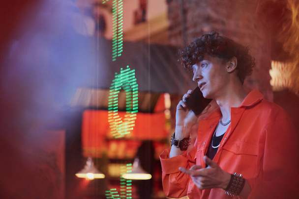 Jongeman met smartphone door oor praten met zijn vriend of vriendin terwijl hij voor de camera tegen de muur in cyberpunk bar of cafe - Foto, afbeelding