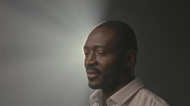 Chest up studio portrait of Black man standing under white spotlight shining from above on graphite background showing palm at camera and smiling - Footage, Video