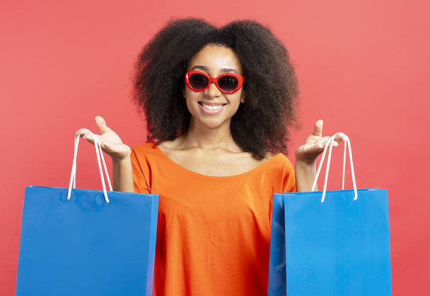  Aranyos, mosolygós nő napszemüvegben, piros háttérrel izolált bevásárlószatyrokkal. Vásárlási koncepció - Fotó, kép
