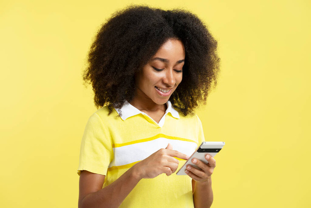 Piękny uśmiech Afroamerykanka trzyma smartfona, za pomocą aplikacji mobilnej zakupy online izolowane na żółtym tle. Szczęśliwa, kręcona hipsterka rozmawiająca na osobności. Koncepcja technologii - Zdjęcie, obraz