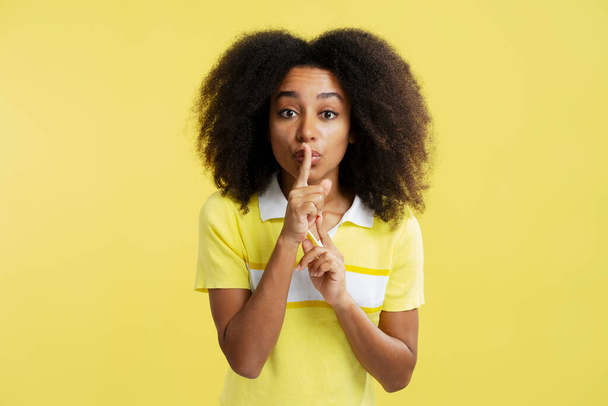 Portret podekscytowanej, uśmiechniętej Afroamerykanki trzymającej palec blisko ust, pokazującej gest milczenia patrzącej na kamerę odizolowaną na żółtym tle. Tajna koncepcja - Zdjęcie, obraz