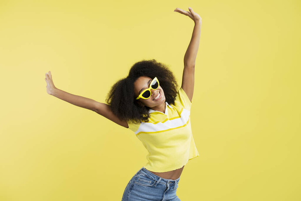 Portrait of friendly positive African American young woman with curly hair wearing stylish yellow sunglasses isolated on yellow background, closeup. Summer concept - Photo, Image