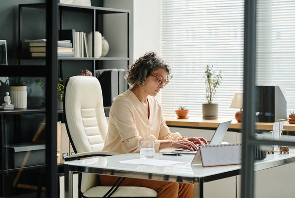 Widok z boku dojrzałej pracodawczyni wpisując na klawiaturze laptopa podczas siedzenia w biurze i pracy z danych online - Zdjęcie, obraz