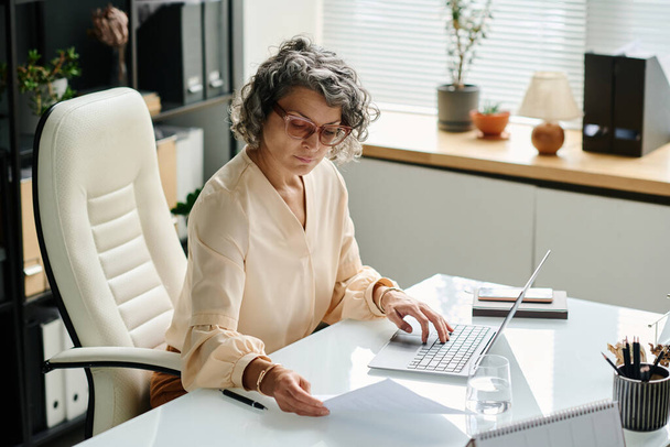 Dojrzałe kobiety pracodawca przeglądając dokument papierowy i wpisując na klawiaturze laptopa przy wprowadzaniu danych osobowych nowych wnioskodawców - Zdjęcie, obraz