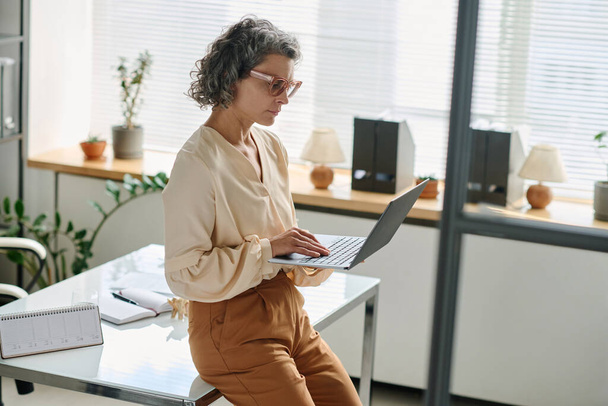 Серйозна зріла жінка-аналітик в тихому розкішному вбранні на клавіатурі ноутбука, сидячи на столі з календарем і відкритим ноутбуком - Фото, зображення