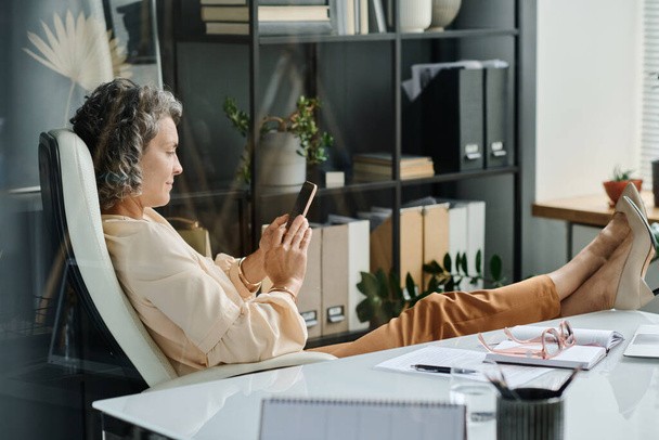 Vista lateral do empresário feminino descontraído maduro sentado em poltrona de couro branco confortável e assistindo vídeo on-line no smartphone - Foto, Imagem