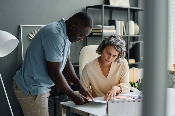 Young confident African American male employee pointing at financial document while explaining its main points to mature female boss - Photo, Image