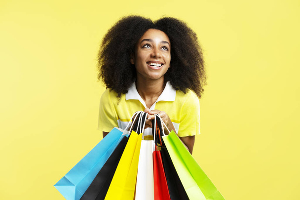 Krásná šťastná Afroameričanka drží barevné nákupní tašky odvrací pohled izolované na žlutém pozadí. Obchod, nakupování, prodej, Black Friday koncept - Fotografie, Obrázek